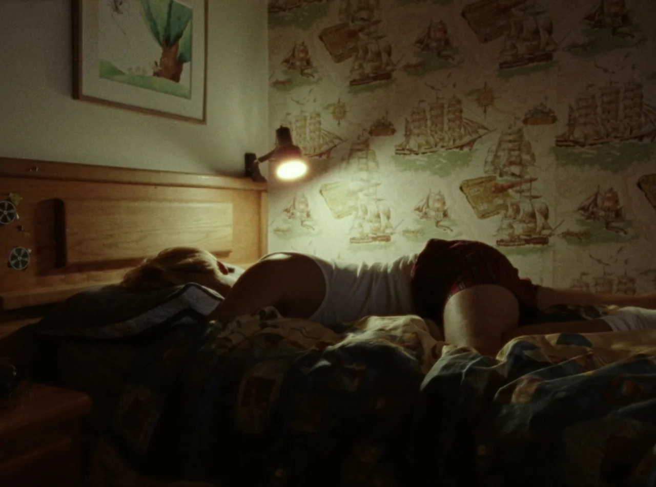 Riceboy Sleeps (2022) movie still: medium — A dimly lit room features patterned wallpaper depicting sailing ships and a wall-mounted lamp. A person…