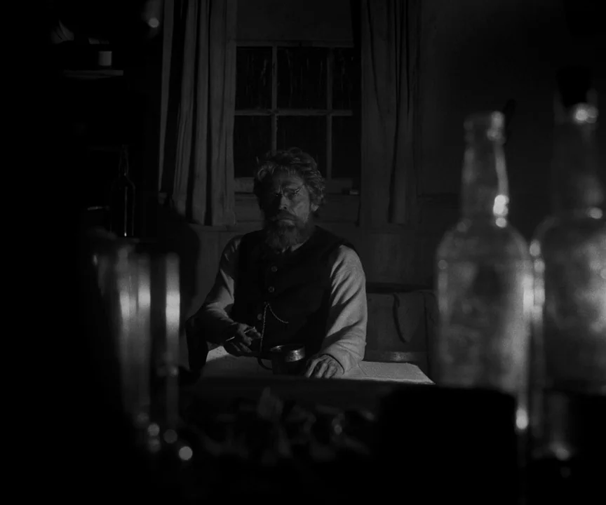 The Lighthouse (2019) movie still: medium — A dimly lit room at night with a small window and dark curtains. An older bearded man sits at a table…