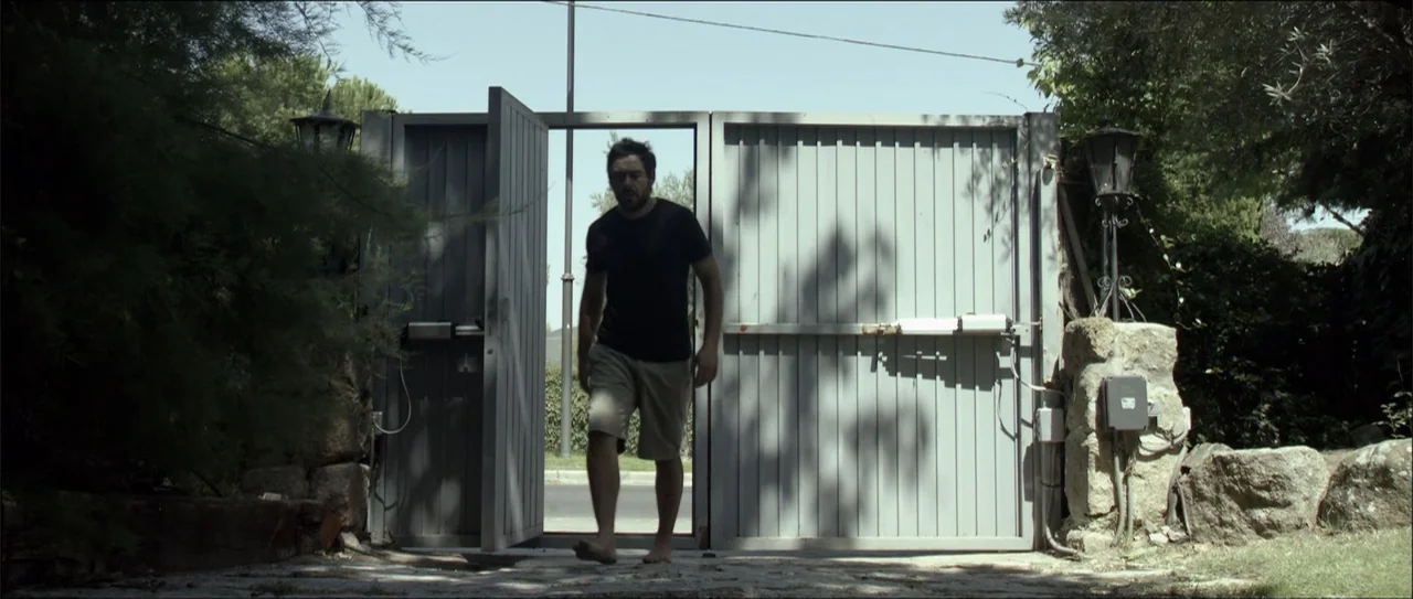Otro verano (2013) movie still: fullbody — A man walks through a light grey metal entrance gate toward the camera. Sunlight casts sharp shadows…