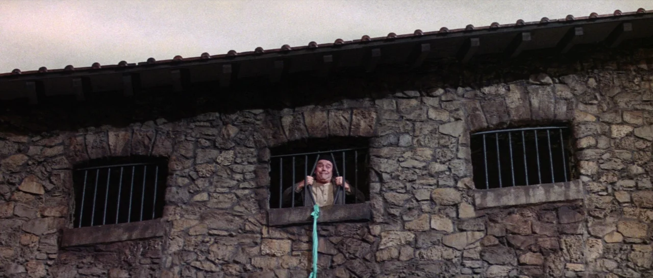 Irma La Douce (1963) movie still: wide — A man looks through a window opening in a rustic, rough-hewn stone wall exterior. He grips two iron bars of…