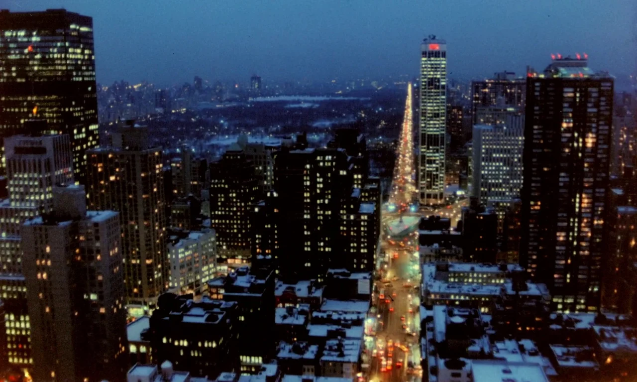 Little Girl Blue (2023) movie still: establishing — An aerial night view shows a dense urban skyline covered in patches of snow. Illuminated…