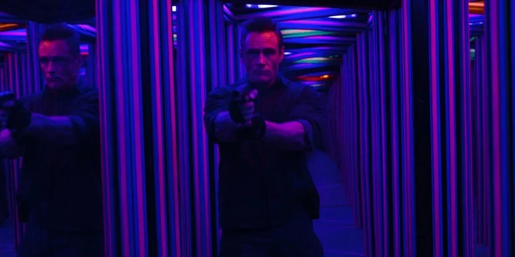 Stranger Things (2019) movie still: medium — A man stands inside a narrow mirror maze corridor illuminated by vibrant neon strips. He points a…