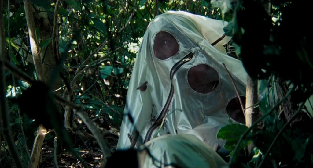 Rescue Dawn movie still: medium — A person wears a metallic shroud while crouched deep within dense, green forest undergrowth. A small snail crawls…