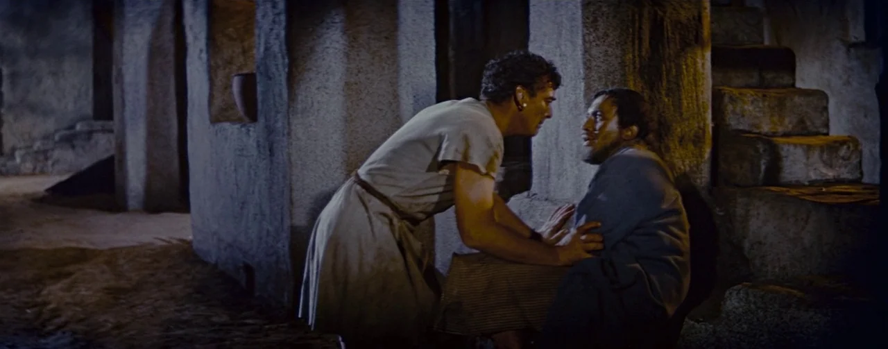 The Robe (1953) movie still: medium — Two men stand within a shadowed stone structure with coarse walls and stairs. A man in a light tunic leans…