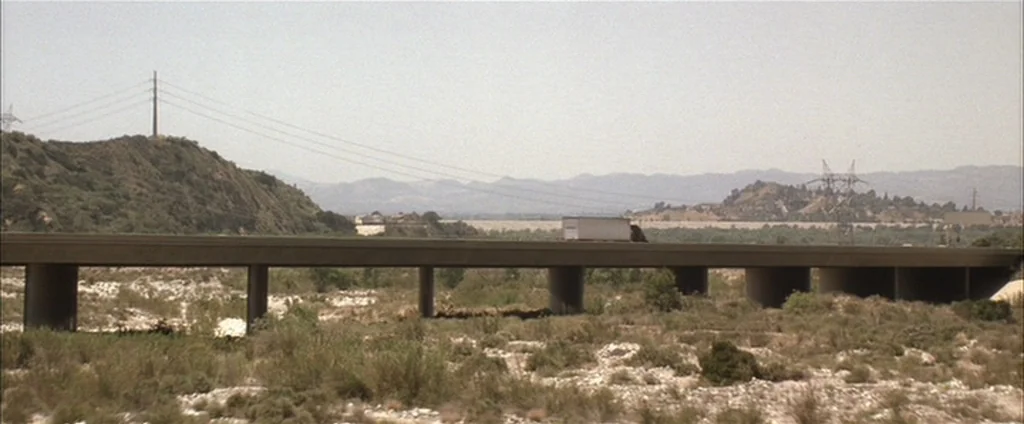 Down In The Valley (2005) movie still: establishing — A long concrete highway overpass spans a dry, brush-filled riverbed under a clear sky. A lone…