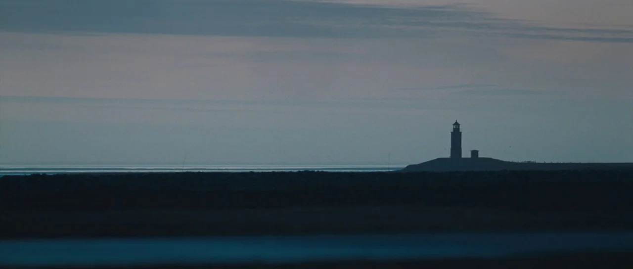 Womb (2010) movie still: establishing — A dark, distant lighthouse stands on a low coastal ridge against a vast sky. The calm, thin line of the…
