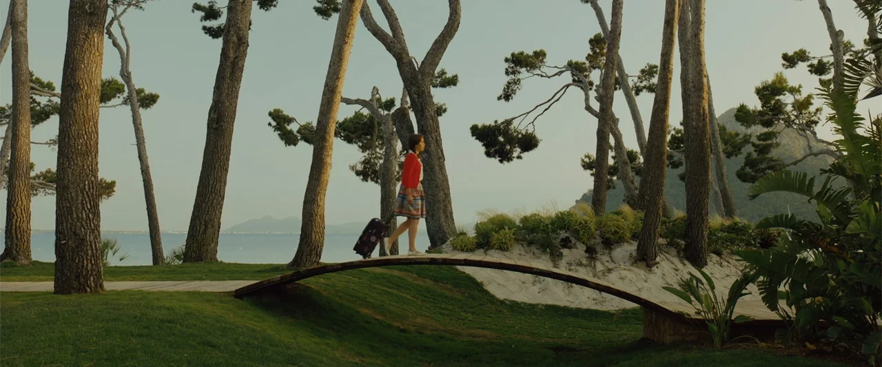 Me Before You (2016) movie still: wide — A coastal park area features tall pine trees and sandy dunes near the sea. A woman in a red cardigan walks…