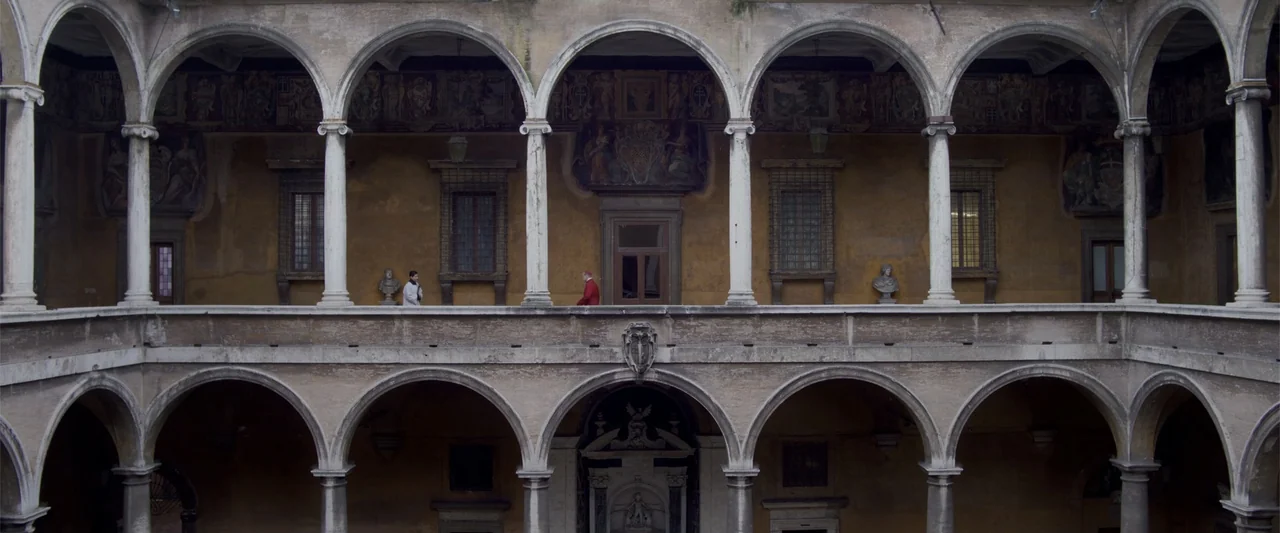 Conclave (2024) movie still: wide — A historic Italian renaissance palace courtyard features layered stone arches and frescoed walls. Two men walk…