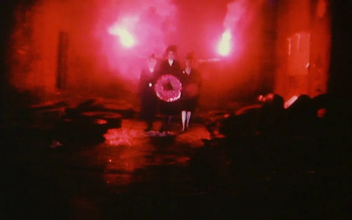The Last of England (1987) movie still: fullbody — A dark outdoor space is illuminated by two bright, smoky red flares. Three individuals stand in a…