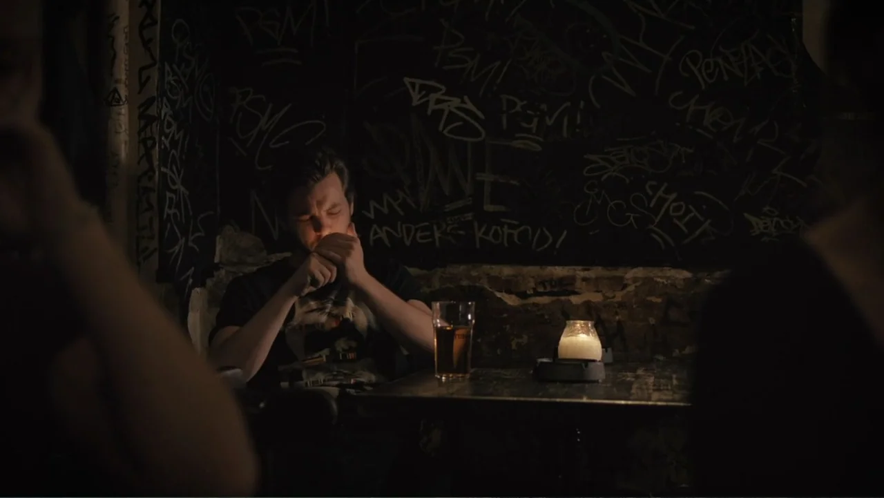 Copenhagen (2002) movie still: medium — A dark, dimly lit bar with graffiti-covered walls and a small wooden table. A man in a dark t-shirt sits…