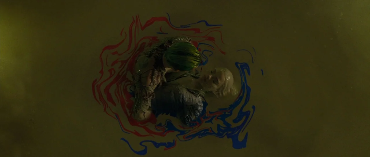 Suicide Squad (2016) movie still: wide — Two individuals are positioned in a large container filled with murky olive-green fluid. They are embraced…