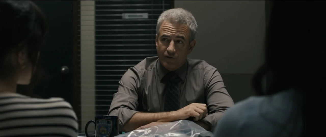 Scream VI (2023) movie still: medium — An interrogation room featuring a dark table and a window with horizontal blinds behind. A middle-aged man in…