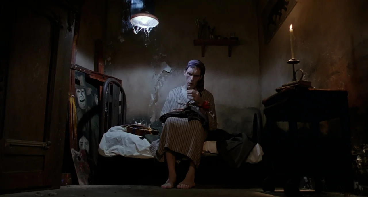 The Addams Family (1991) movie still: medium — A cramped, dilapidated room features peeling walls and a single bed. A pale man sits on the bed…
