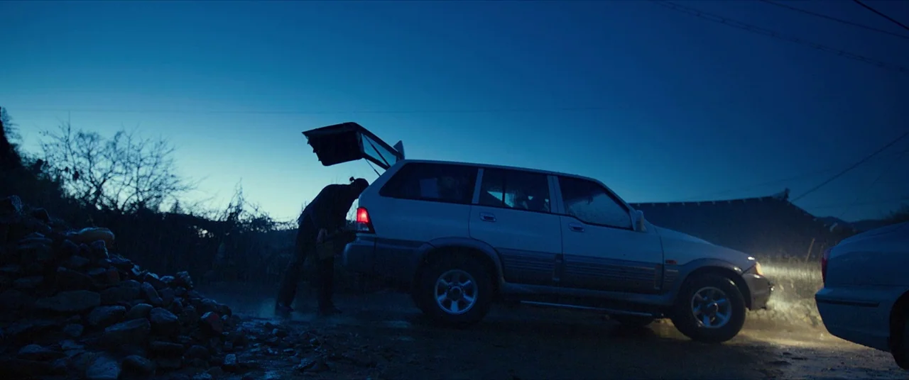 The Wailing (2016) movie still: wide — A rural road at blue hour features a silver SUV with its trunk open. A man stands beside the vehicle,…