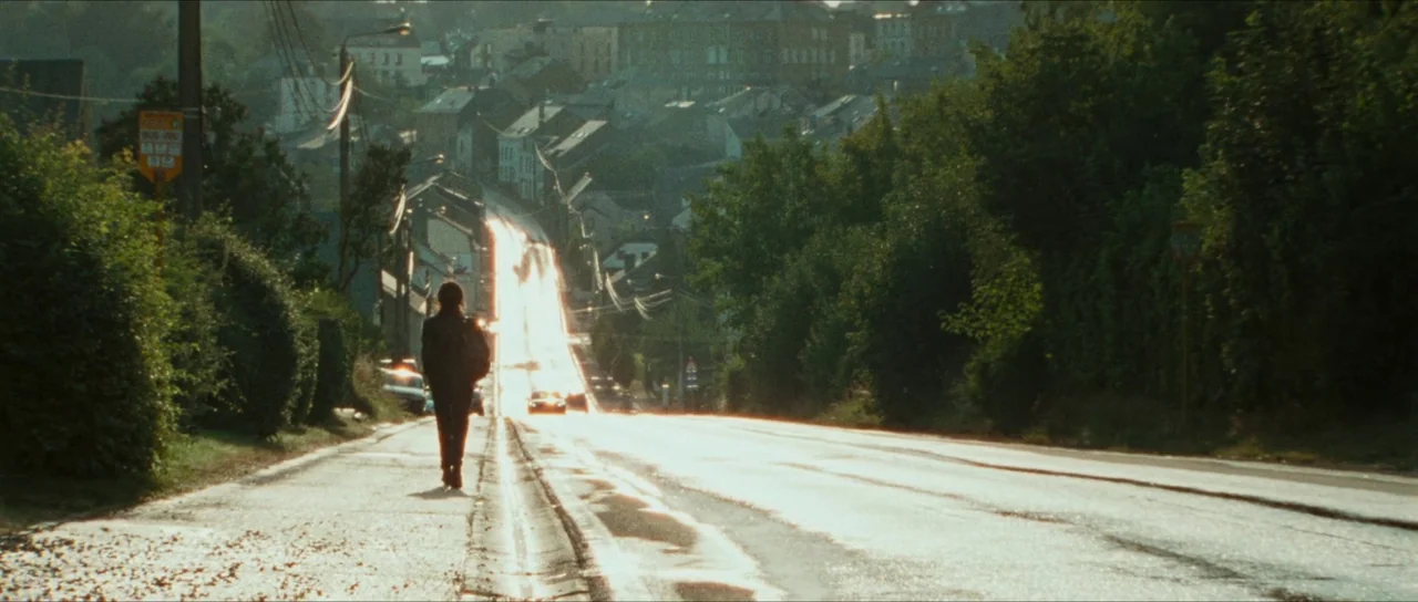 Inexorable (2021) movie still: wide — A long paved road leads uphill toward a European-style town with dense trees bordering it. A single person…