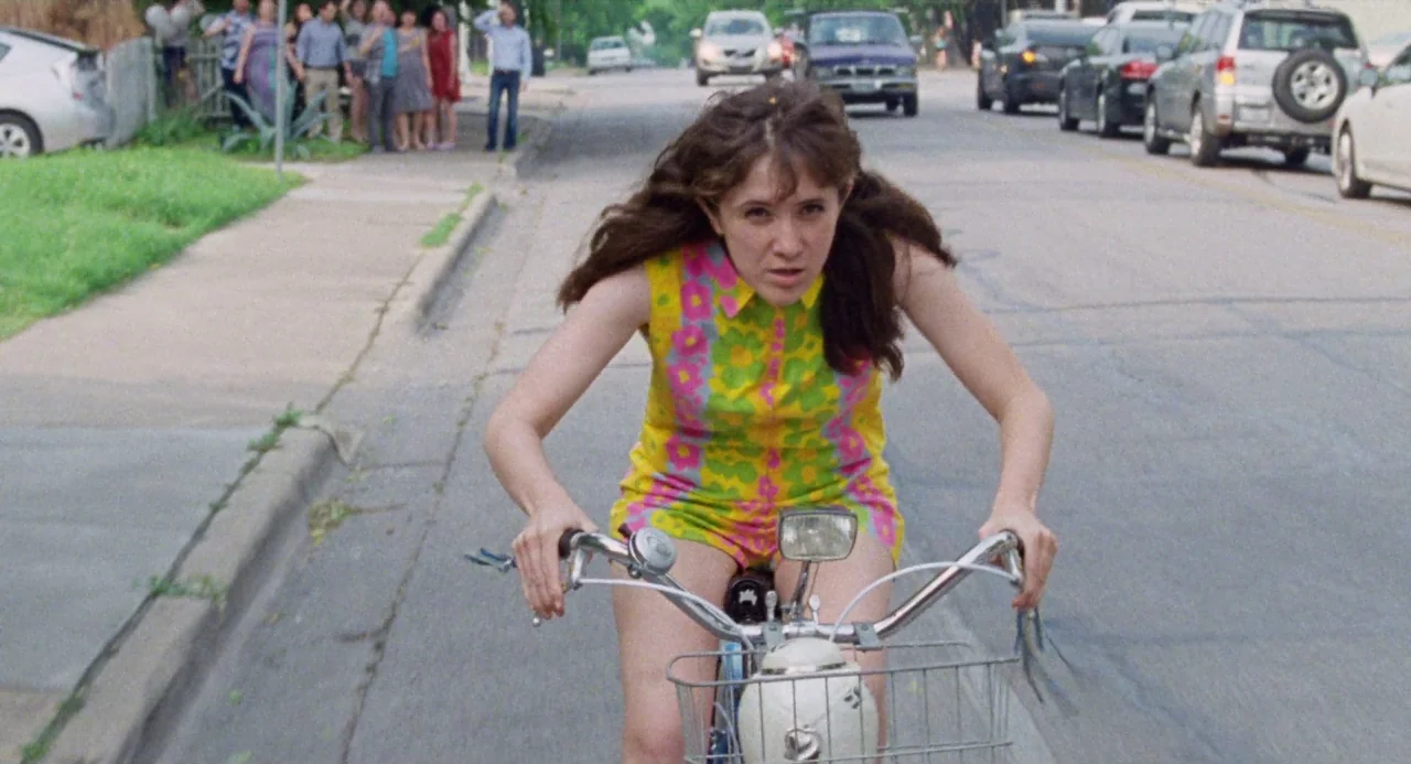 Mr. Roosevelt (2017) movie still: medium — A suburban residential street during the day with cars parked along the curb. A young woman in a brightly…