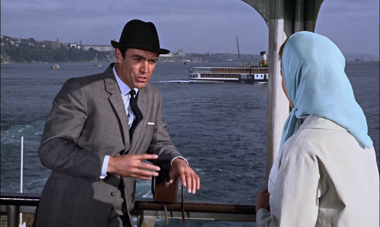 From Russia With Love (1963) movie still: medium — A man in a suit and a woman in a headscarf stand on a deck. The background shows a passenger boat…