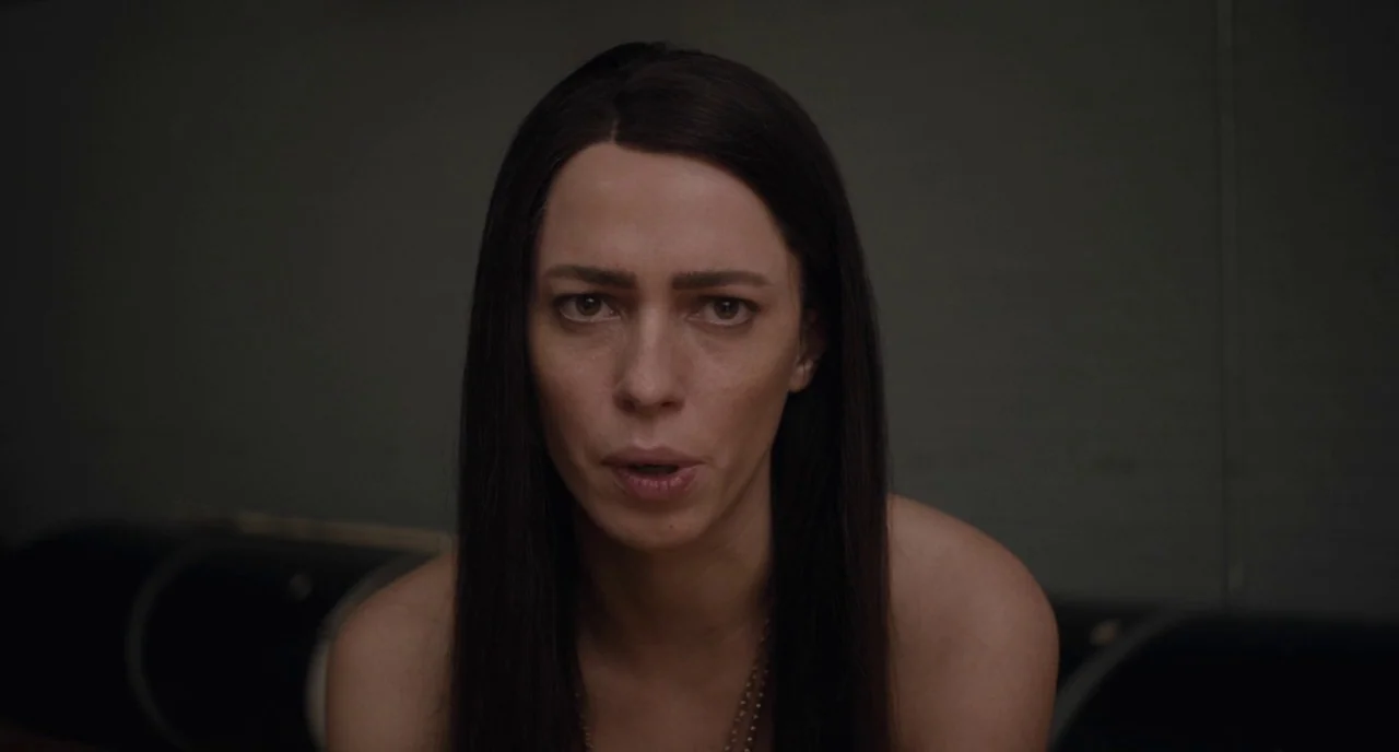 Christine (2016) movie still: closeup — A dimly lit room with plain dark walls serves as the backdrop. A woman with long dark hair gazes directly…