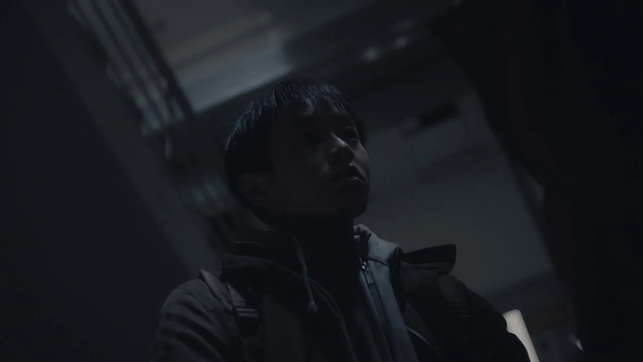 An Elephant Sitting Still (2018) movie still: medium — A dimly lit industrial stairwell with metal railing visible in the shadows. A young man…