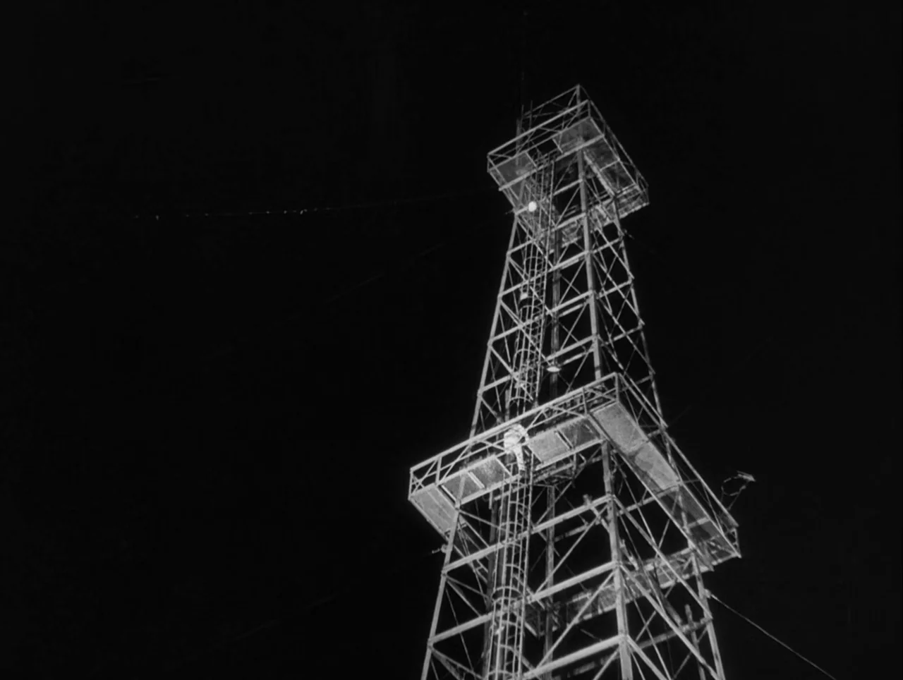 The Wages of Fear (1953) movie still: wide — A tall iron oil derrick stands in darkness against a pitch-black night sky. The structure is…