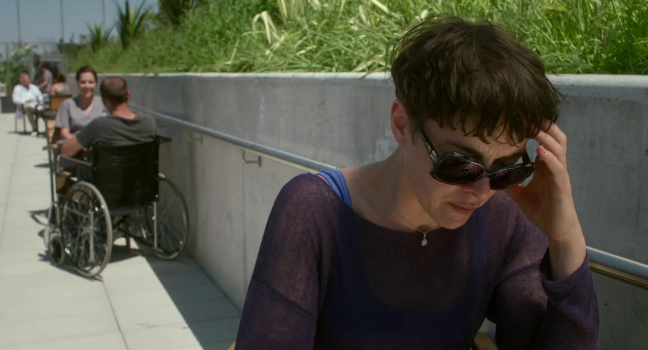 Maps to the Stars (2014) movie still: medium — A modern outdoor terrace features a long concrete wall with tall grasses behind it. A woman sits in…
