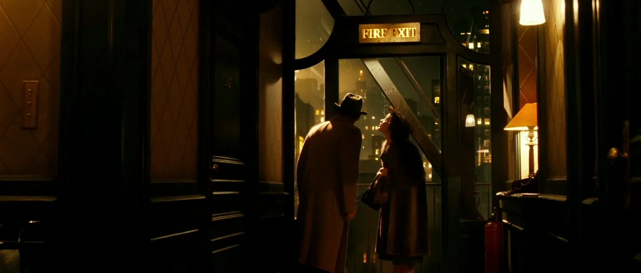 La Vie En Rose (2007) movie still: medium — An interior hallway leading to a glass fire exit door at night. A man and a woman stand together facing…