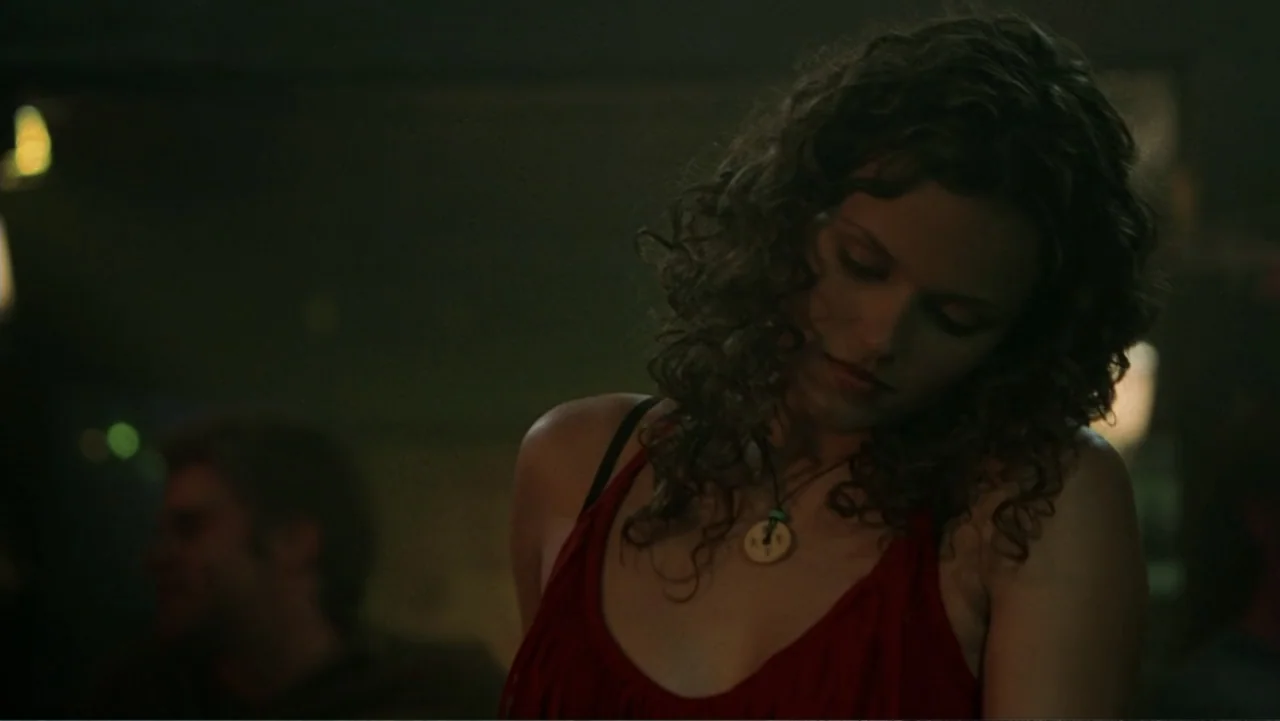 The Butterfly Effect (2004) movie still: medium — A dimly lit indoor space with blurred figures in the background. A woman in a red top stands in…