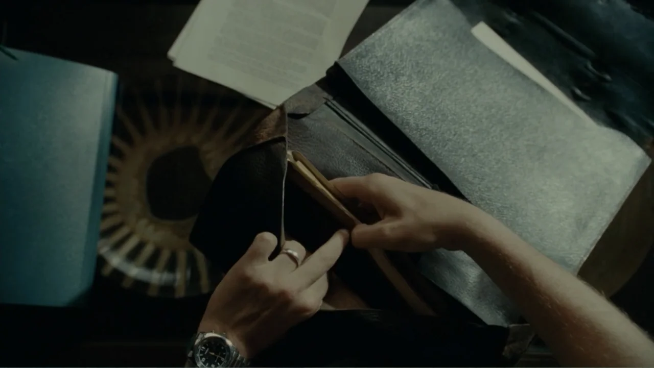 The Words (2012) movie still: closeup — A dark wooden surface contains a leather briefcase, loose documents, and a round patterned coaster. Two…