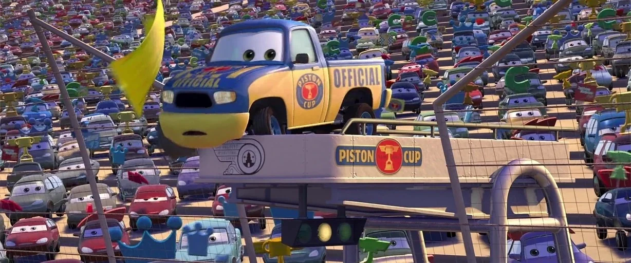 Cars (2006) movie still: wide — A crowded racetrack grandstand is filled with many diverse anthropomorphic cars. An official blue and yellow truck…