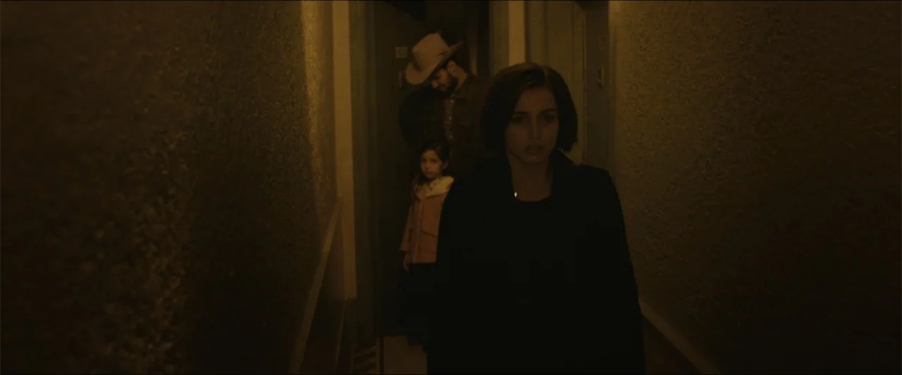 Exposed (2016) movie still: medium — A dimly lit narrow hallway with textured walls features a woman walking forward. Behind her stand a man wearing…