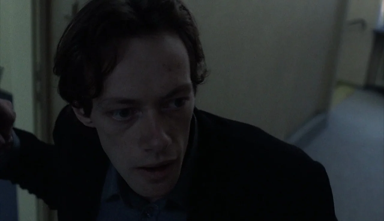 Angst (1983) movie still: closeup — A dimly lit indoor hallway with plain walls and low visibility. A man stands in the shadow looking to his side…