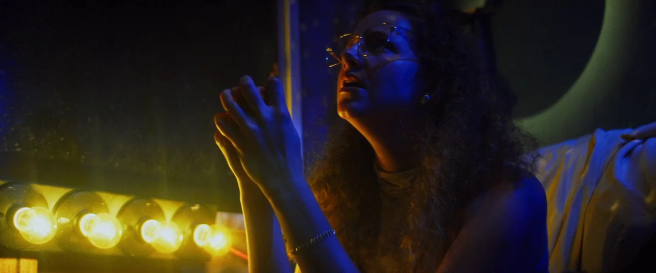 Revealer (2022) movie still: closeup — A dark, cramped dressing room features a vanity with a row of illuminated bulbs. A woman in glasses gazes…