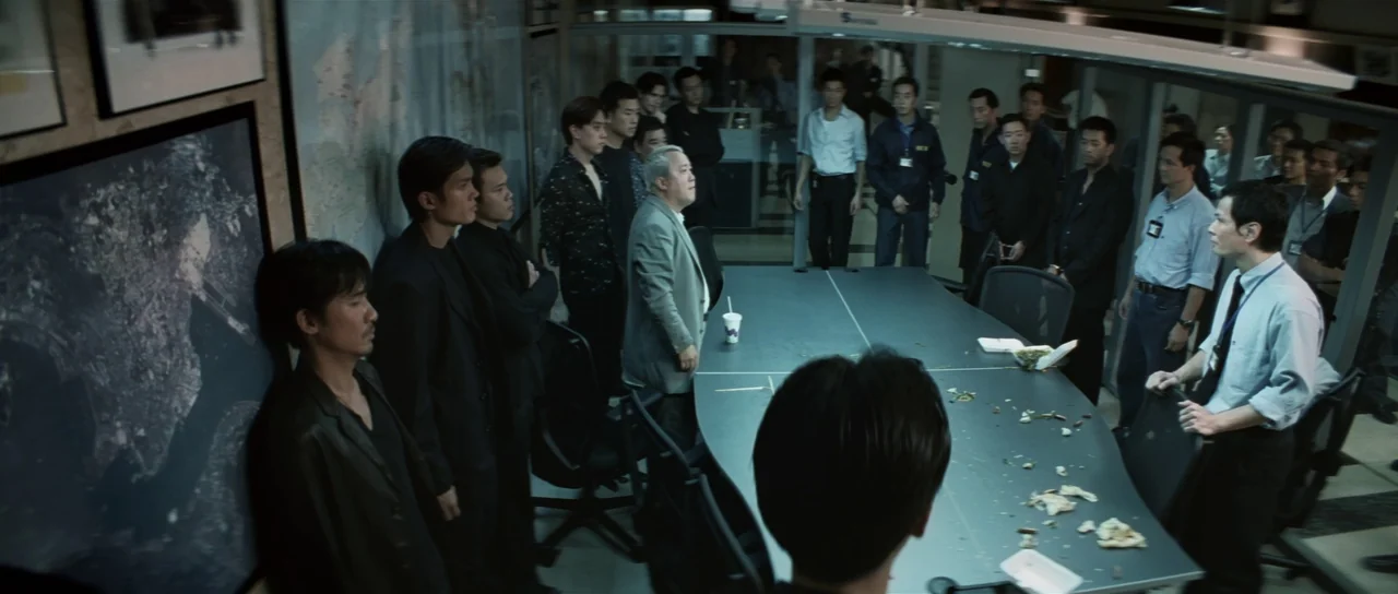 Infernal Affairs (2002) movie still: wide — A modern, glass-walled conference room contains a large oval table with scattered food waste. Numerous…
