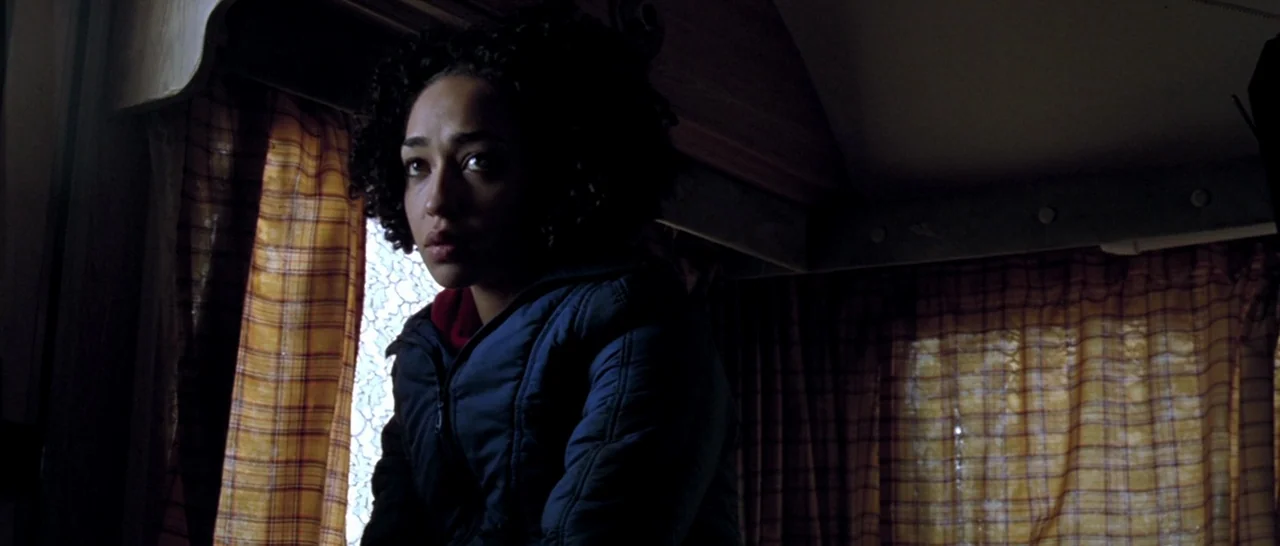 Isolation (2005) movie still: medium — A dark, cramped caravan interior is lined with vintage-style plaid curtains. A young woman sits in the dim…