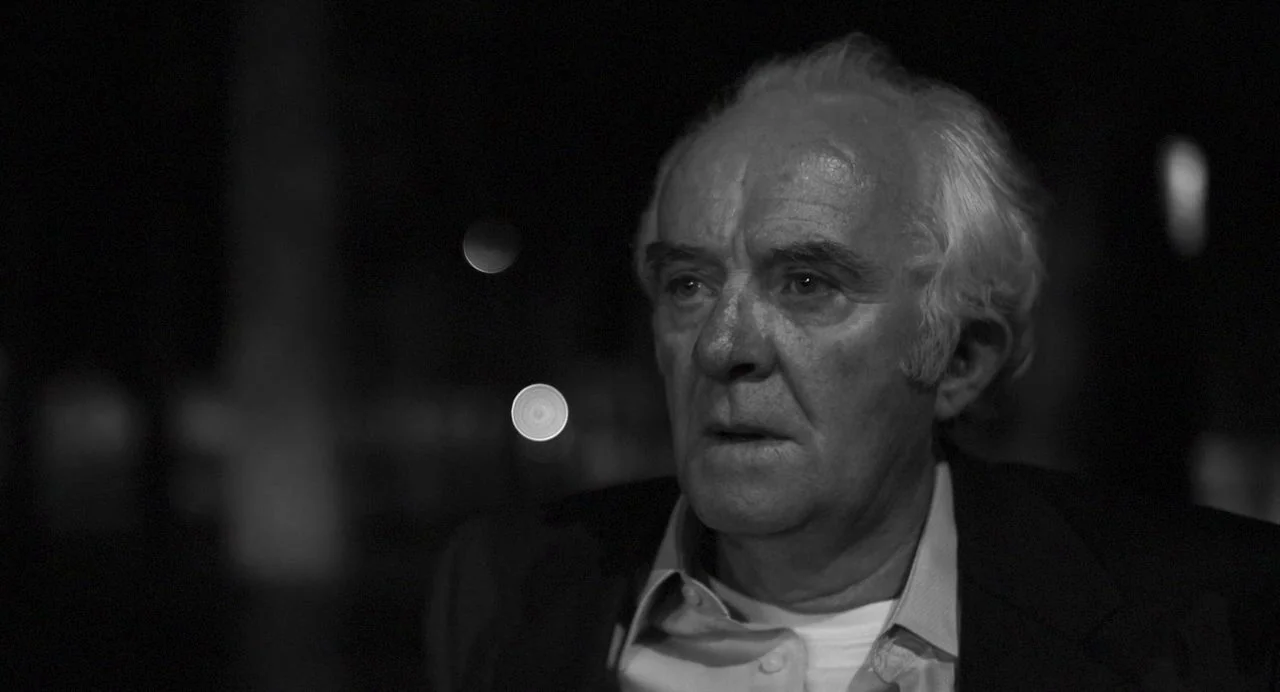 Song of Granite (2017) movie still: closeup — A dark outdoor space with bokeh light points blurred into the background. An elderly man in a suit…