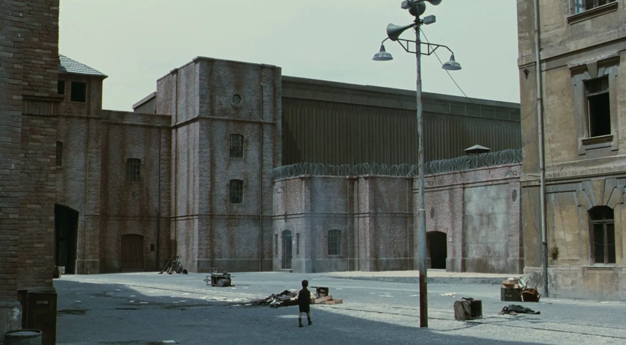 Life is Beautiful (1997) movie still: establishing — A wide, austere courtyard is enclosed by high brick walls topped with barbed wire. A young…