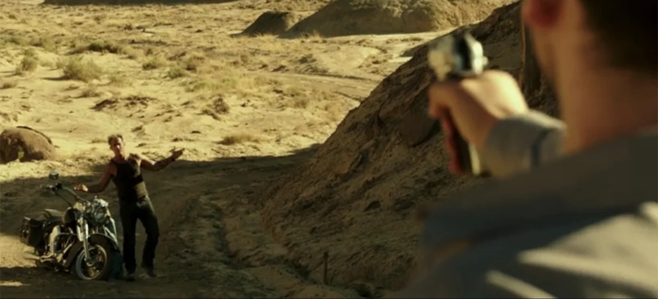 Blood Father (2016) movie still: wide — A dry desert landscape features a man standing beside a parked heavy motorcycle. A person in the foreground…