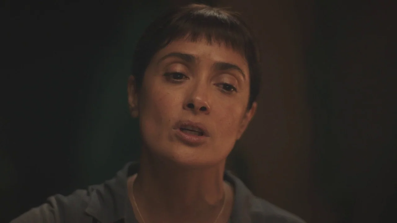 Beatriz at Dinner (2017) movie still: closeup — A dimly lit interior space with a dark, indistinct background. A woman looks slightly off-camera…