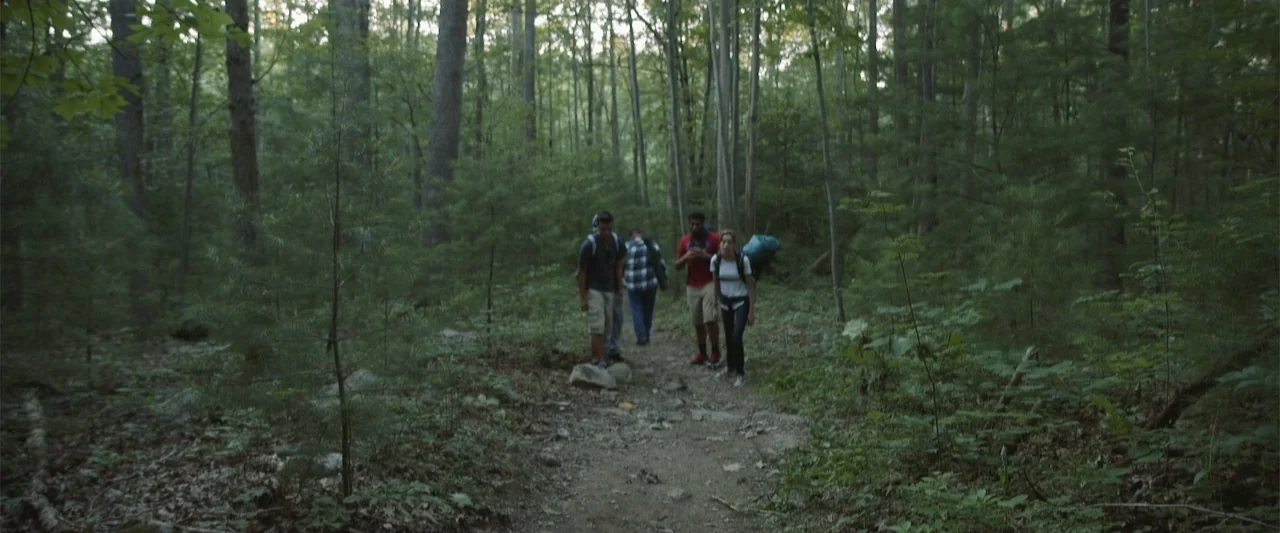Darkness Waits (2020) movie still: wide — A group of four hikers walks along a dirt trail deep within a forest. They carry backpacks while…