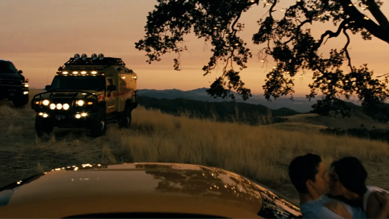 Transformers (2007) movie still: wide — A grassy hilltop at sunset features an off-road utility vehicle parked nearby. A man and woman embrace and…