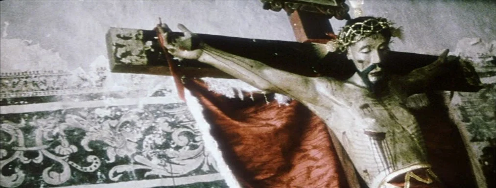 Japón (2002) movie still: medium — A wooden statue of a crucified figure hangs against a wall with ornate painted motifs. The sculpture features a…