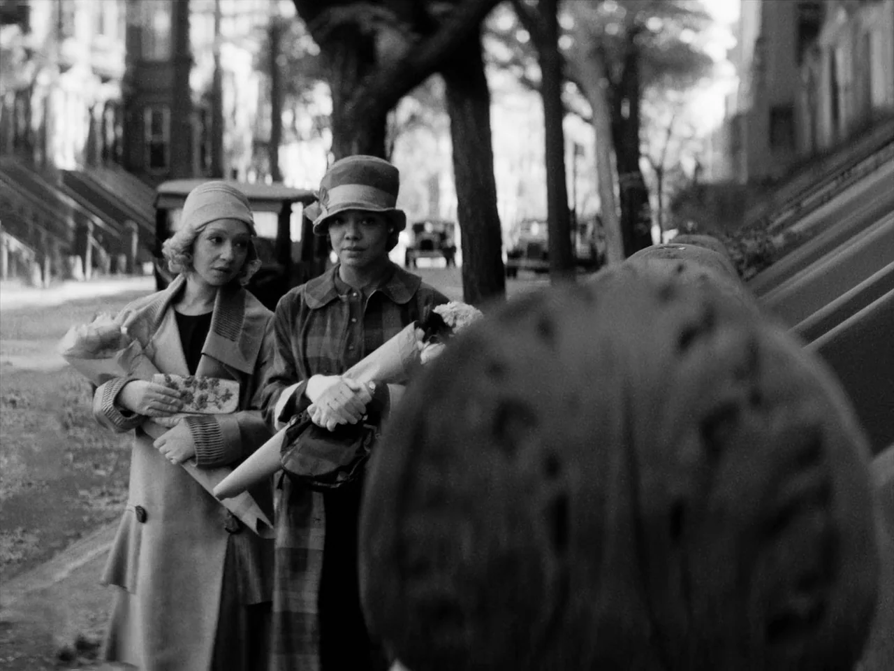 Passing (2021) movie still: medium — Two women walk along a sidewalk past trees and period architecture in black and white. They carry bouquets of…