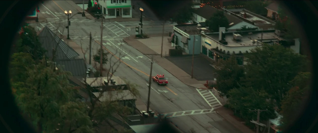 White Noise (2022) movie still: wide — A quiet town intersection is viewed through an obscured, dark circular frame or lens. A single red vehicle…