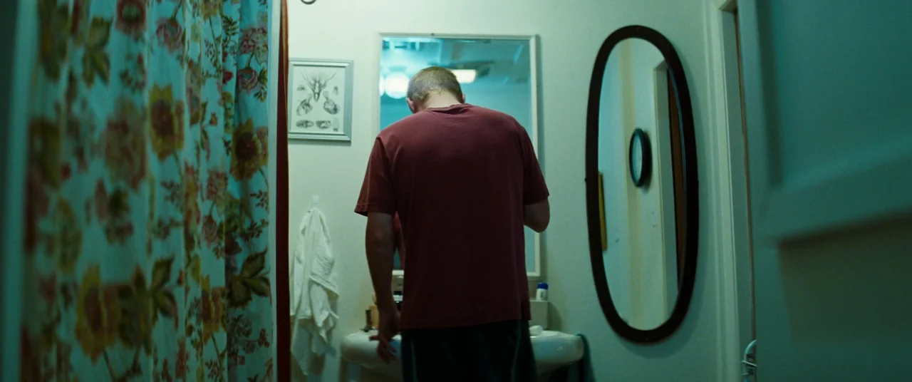Kinds of Kindness (2024) movie still: medium — A small bathroom features a floral shower curtain and a wall-mounted oval mirror. A man with short…