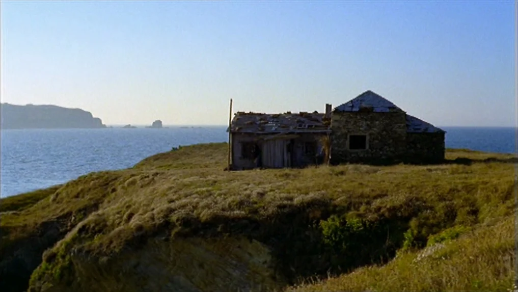 The Scarlet Letter (1973) movie still: establishing — A weathered stone cottage sits atop a grassy coastal headland overlooking the sea. The horizon…