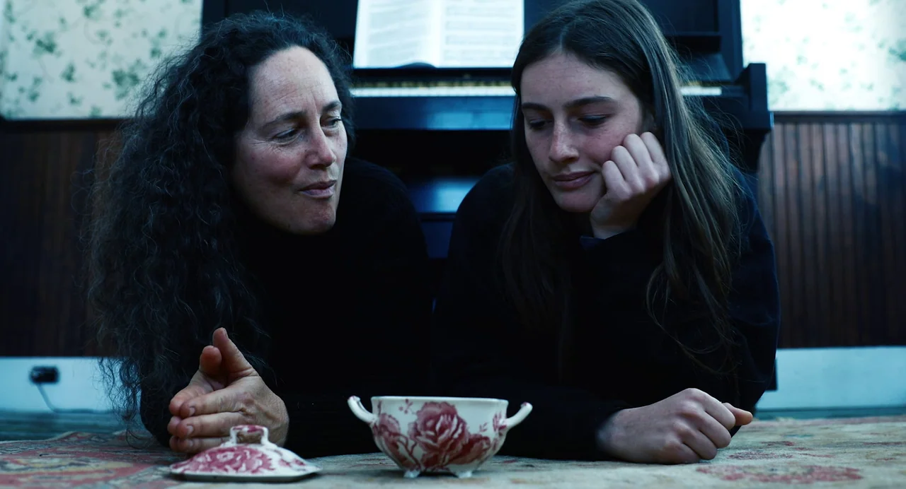 Hellbender (2021) movie still: medium — A residential room features a dark wood-paneled wall and a piano in the background. Two women lie prone on a…