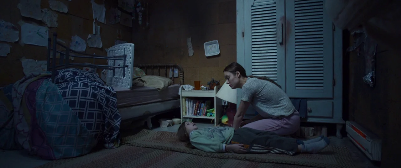 Room (2015) movie still: wide — A dimly lit room with walls covered in children's drawings and a wooden armoire. A young woman kneels on the floor…