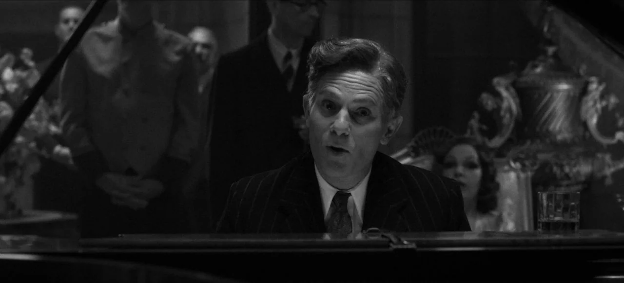 Mank (2020) movie still: medium — A dimly lit ballroom interior with figures in the background. A man sits at a grand piano looking forward.