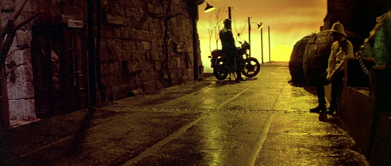 Querelle (1982) movie still: wide — A stone pier stretches toward a setting sun during a vivid golden hour. A man sits on a low wall while a…