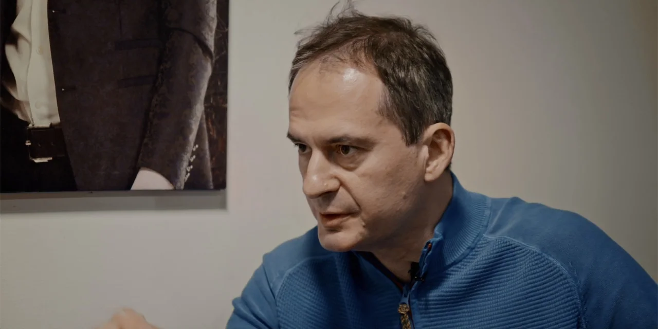 Navalny (2022) movie still: closeup — A plain interior room features a neutral wall decorated with a framed photograph. A man in a blue sweater…